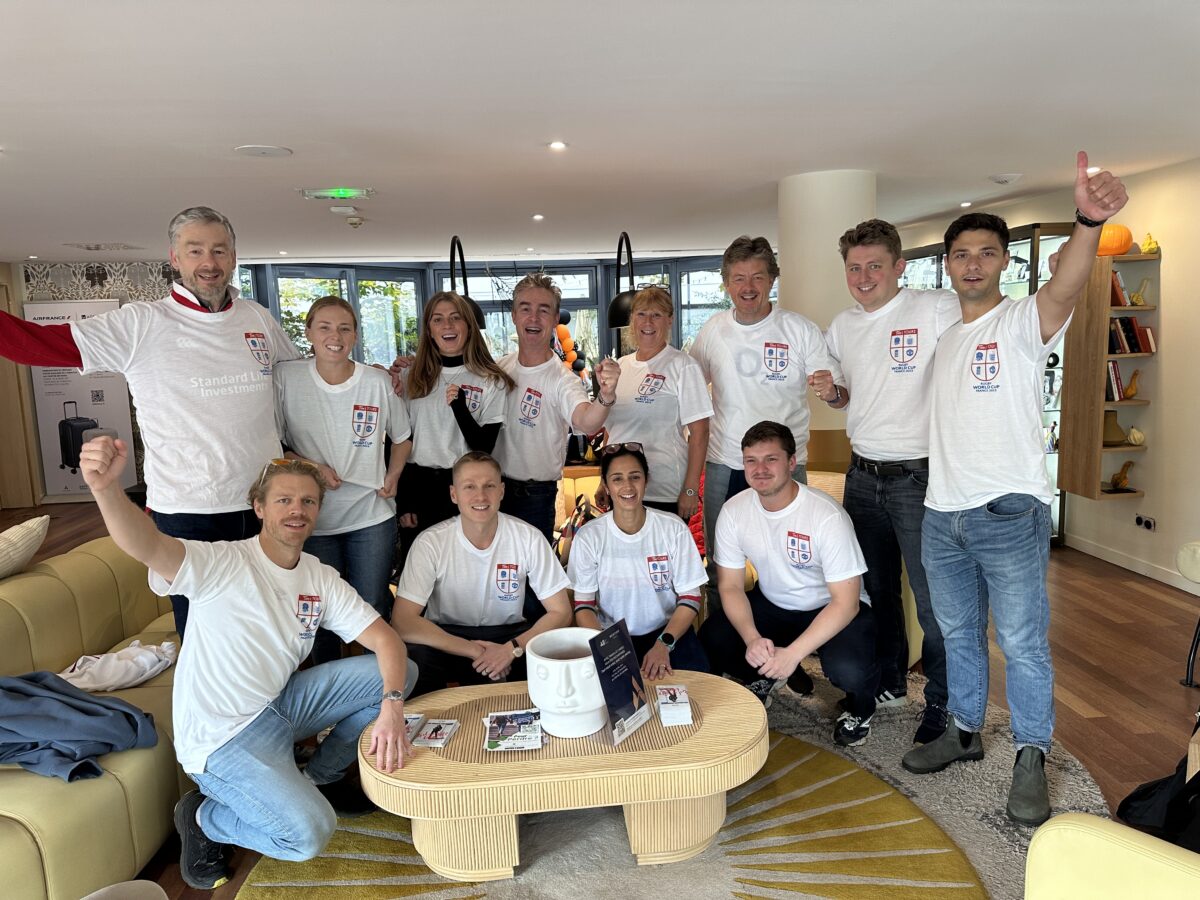 A group of 12 people of different ages wearing white T shirts. There are 8 men and 12 women of different ages in two rows. The back row of 8 are all standing up and the front row of 4 are squatting down.