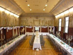 A hall ready for an event with three long tables covered with white tablecloths and a line of green seats on each long side of the table. Around the room are painted murals showing soldiers.
