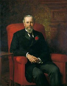 Portrait of a man sitting down. He is wearing a suit and has grey hair and a beard.